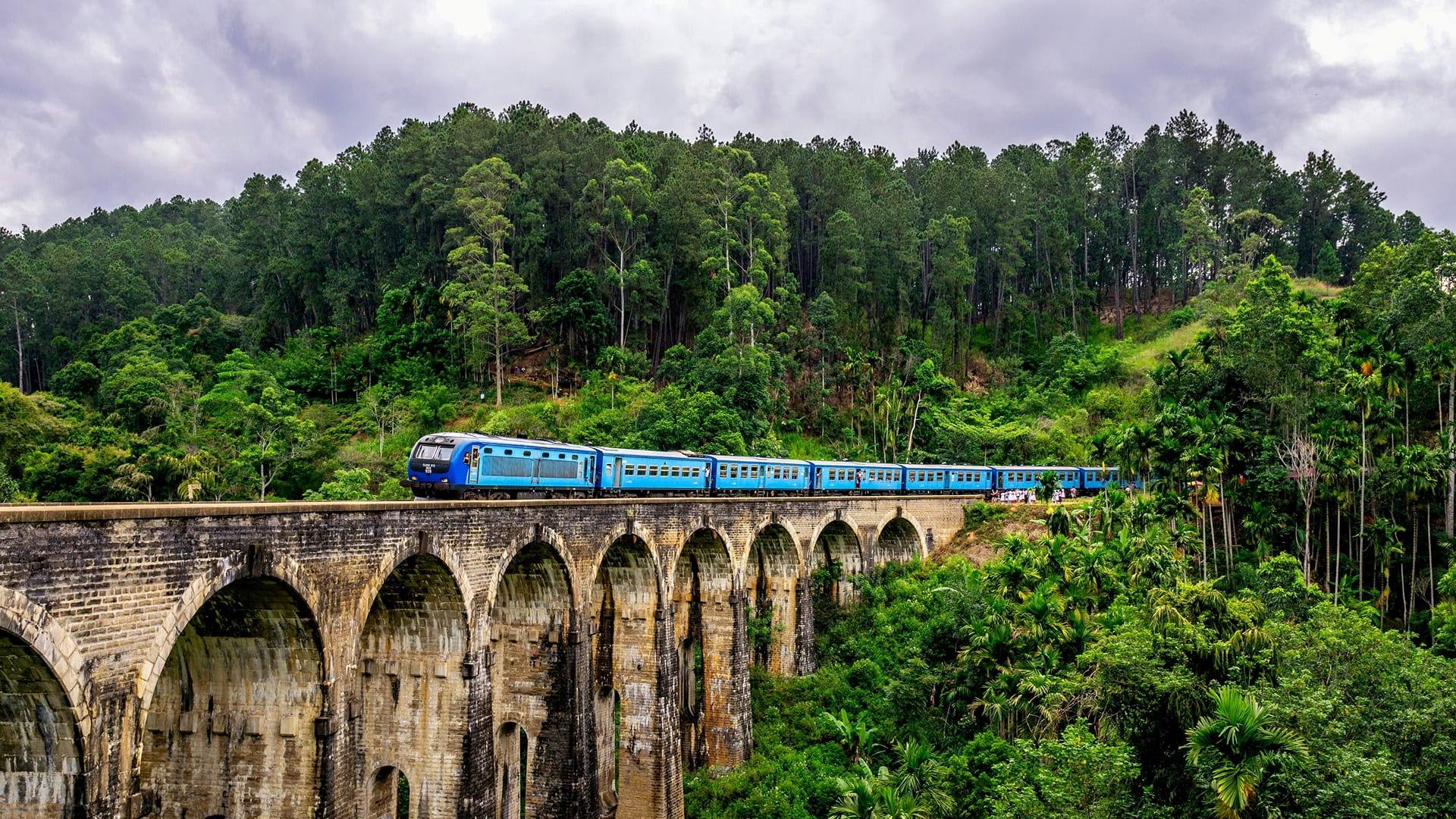 A bright blue train travelling atop the nine arch bridge in Ella