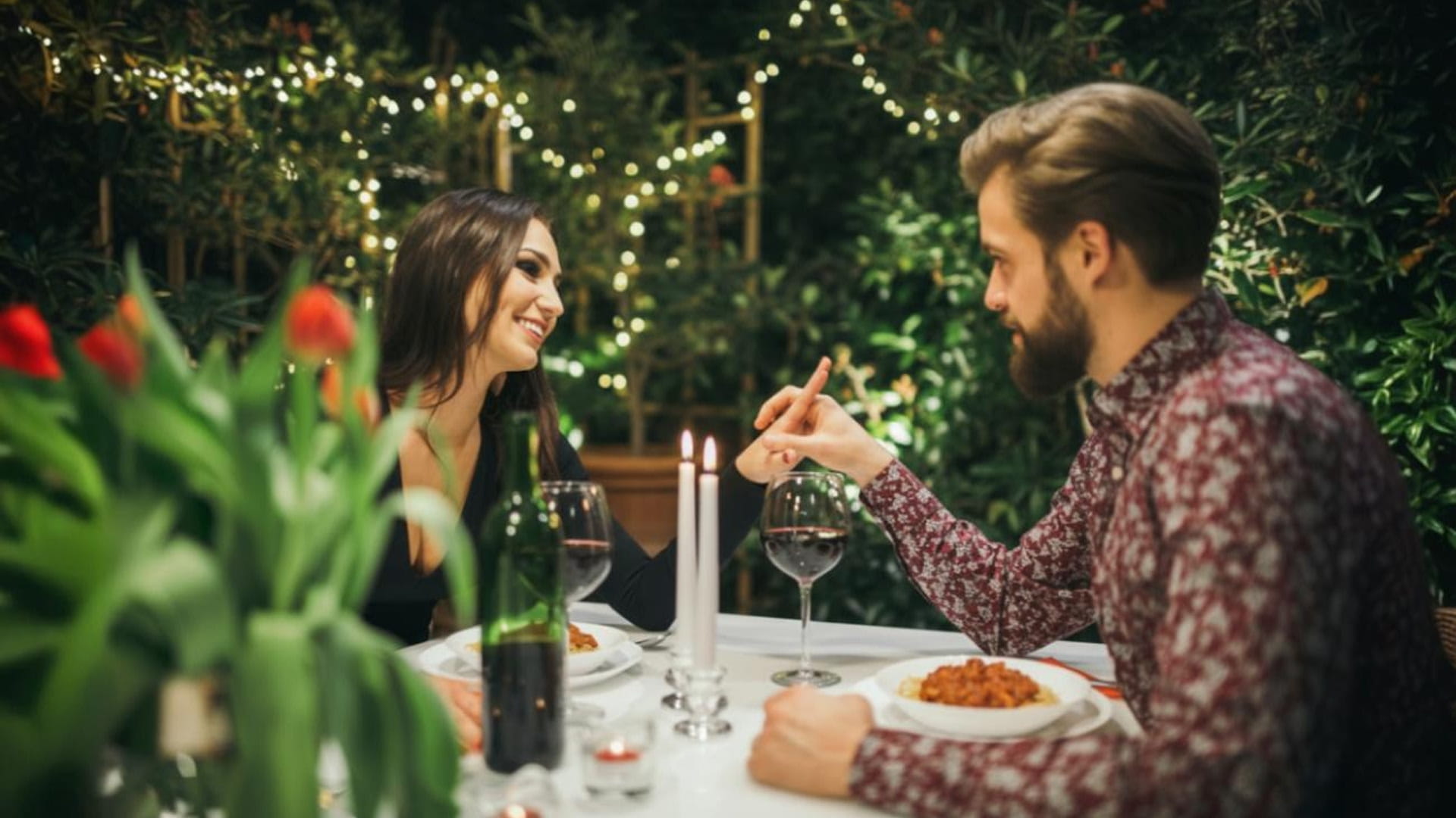 A couple holding hands at a romantic dinner with fairy lights surrounding the area